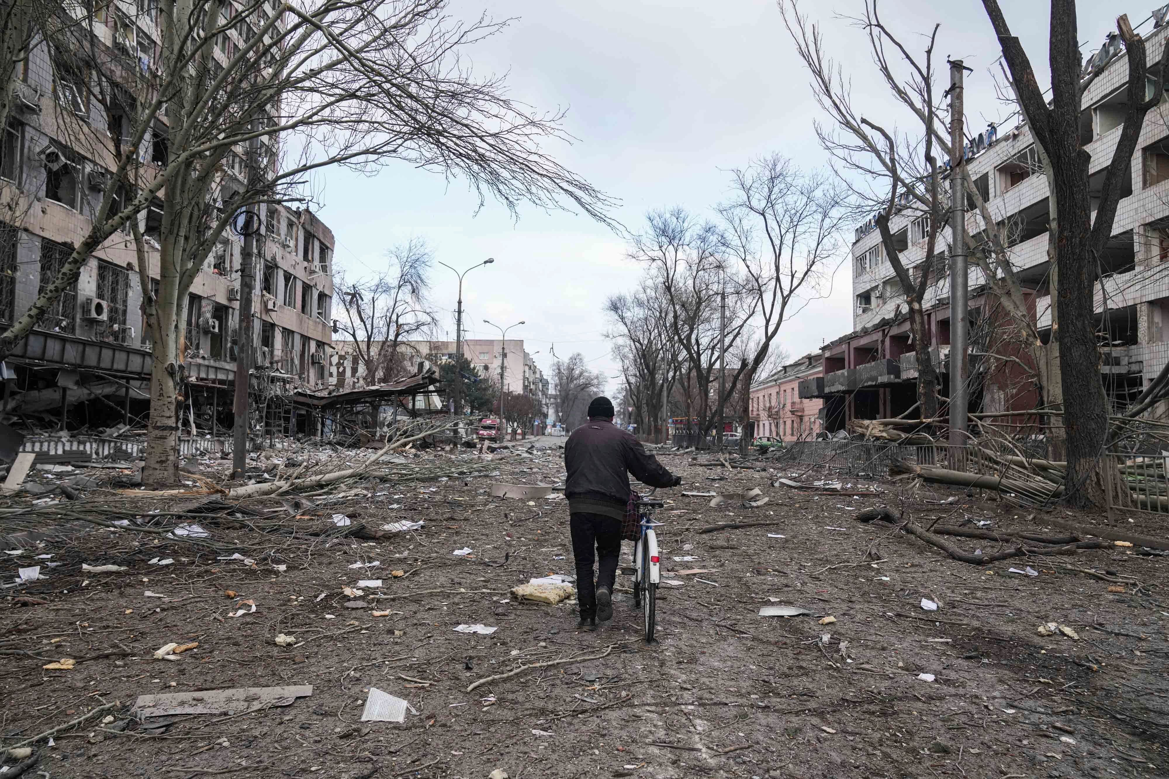 A cyclist walks down a damaged road with destroyed buildings on either side