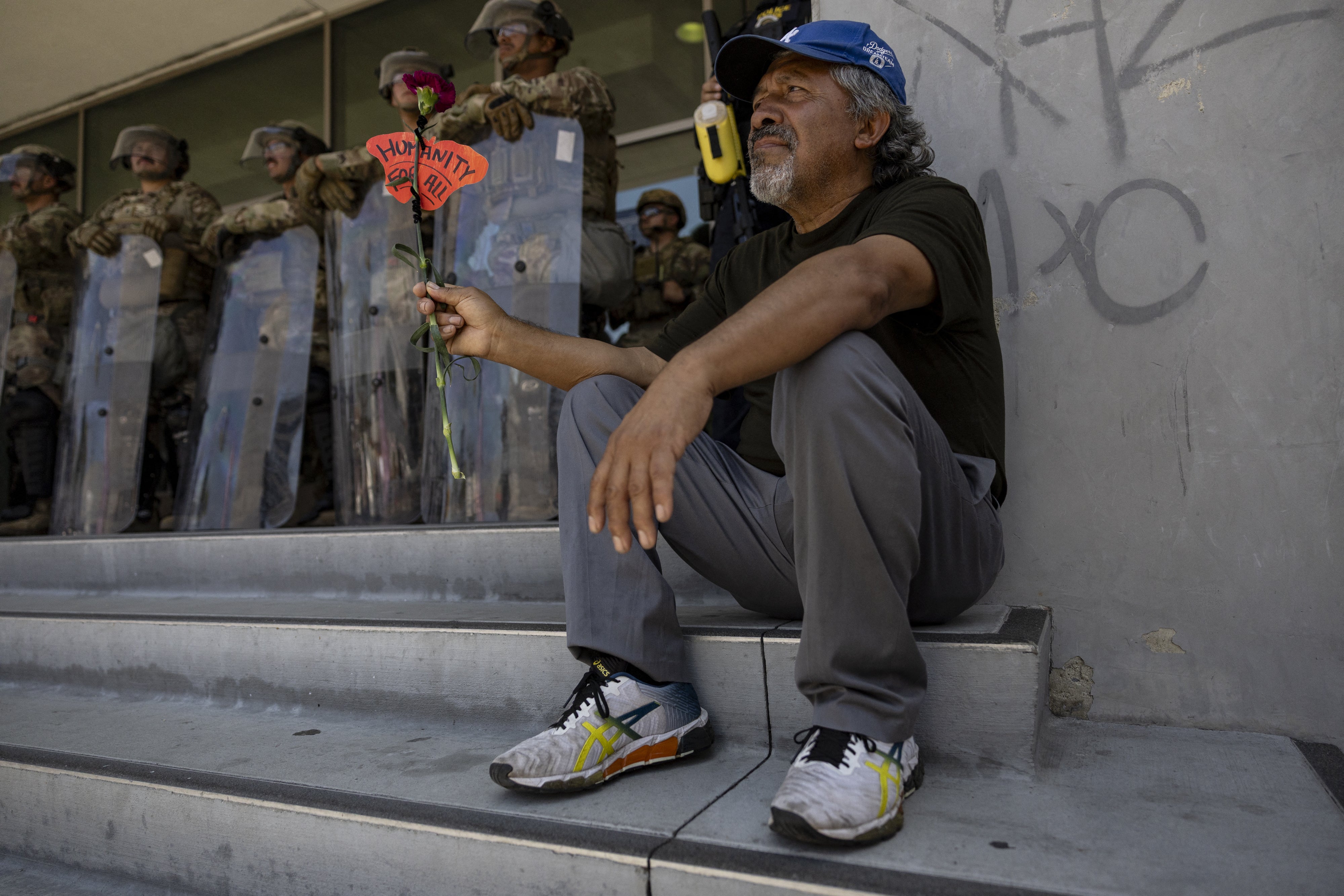 A man holds a flower and the message "Humanity for All" in front of a line of soldiers