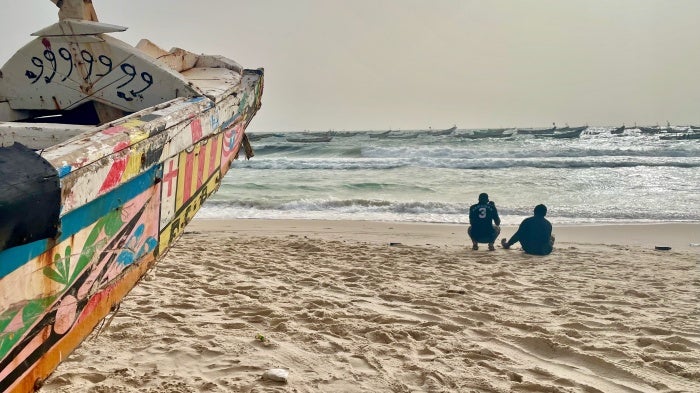 Two men sitting on a beach near wooden boats