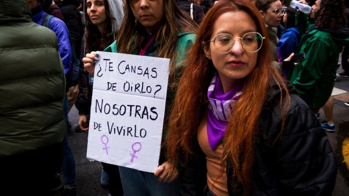 Two women hold a sign that reads “You’re tired of hearing it? We're tired of living it.”