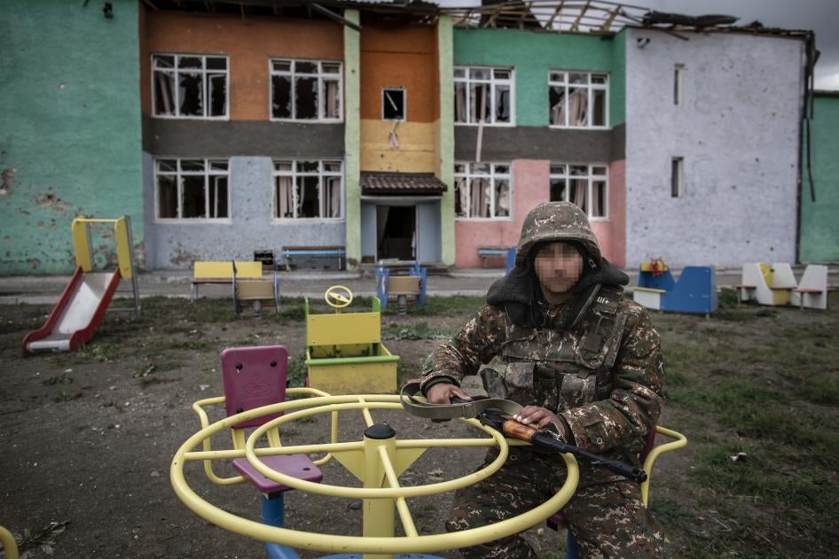 A soldier sits at playground equipment in front of a damaged school building