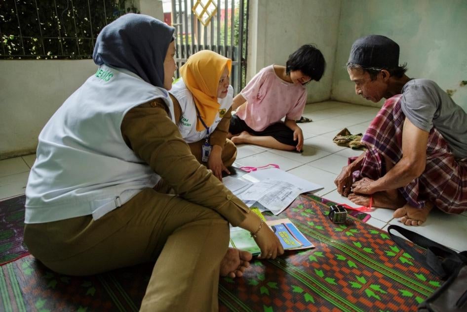 Two workers sitting on the floor with a man and a girl, showing them leaflets spread on the floor.