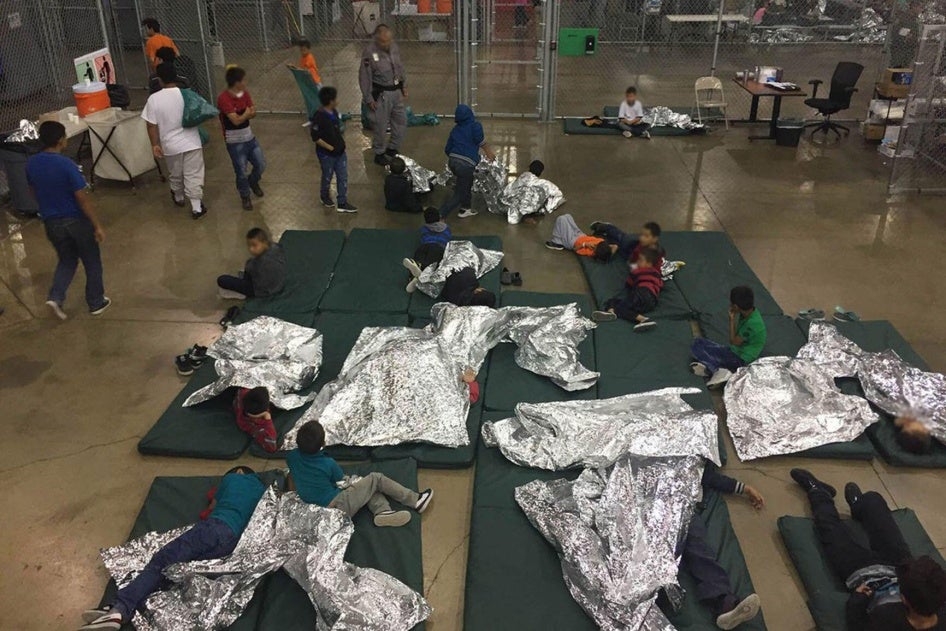 Children in a large indoor fenced-in holding area, wrapped in metallic blankets