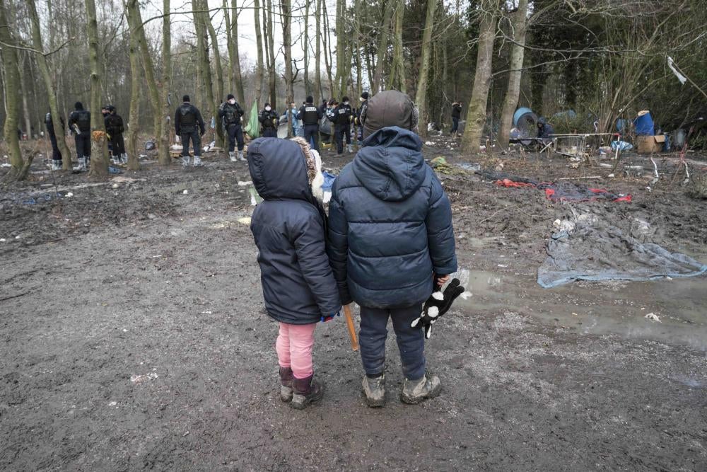 Two children wearing winter coats stand in a forest in front of a group of police officers