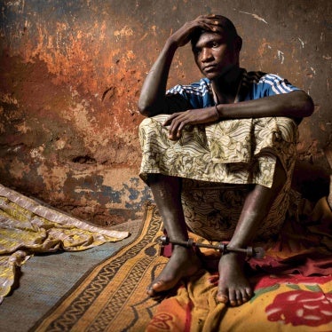 A man sitting on the floor in a dilapidated room, with his ankles locked by a metal bar.